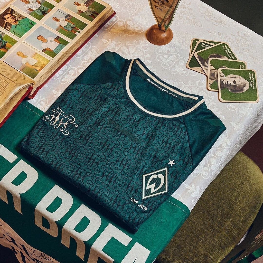 vA17ggJ4nNJBg1j | UAE SPORT STORE Teal soccer jersey with a patterned front, club crest and a star, folded on a table with memorabilia cards nearby.