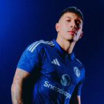 Portrait of a man in a blue Manchester United jersey posing against a dark blue background with a focused expression.