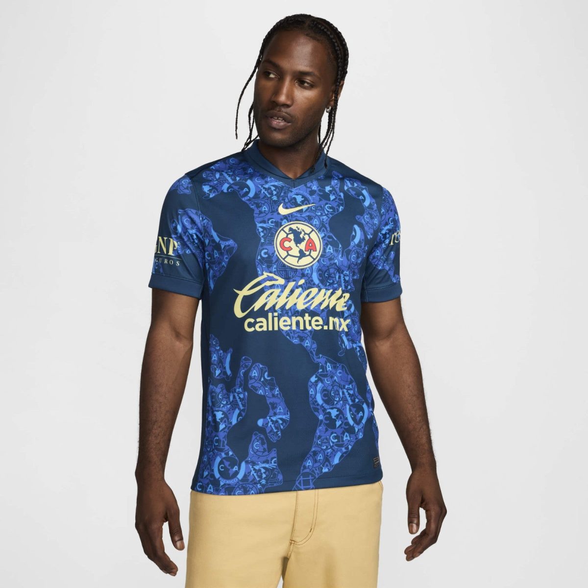 Man wearing a blue patterned football jersey with a yellow CALIENTE sponsor logo and world globe crest on the chest, standing against a light background.
