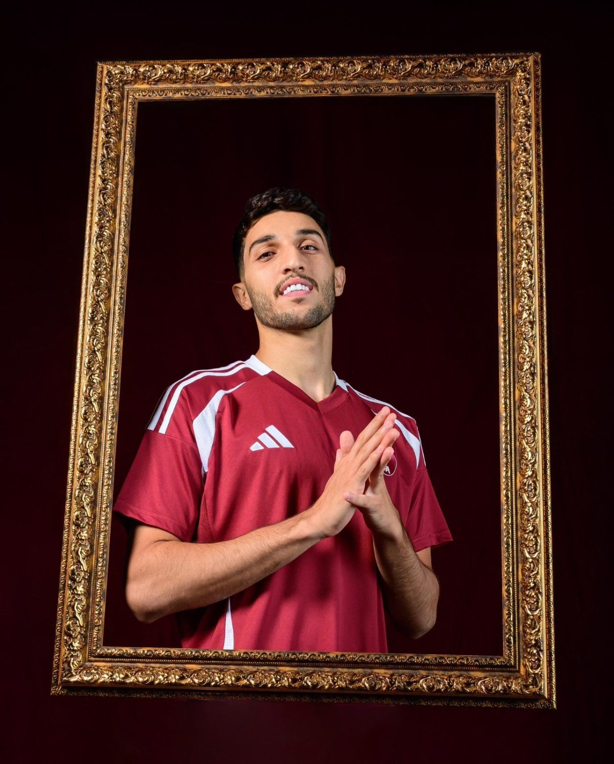 Man in a maroon Adidas jersey stands inside an ornate gold frame, hands pressed together in prayer against a dark burgundy background.
