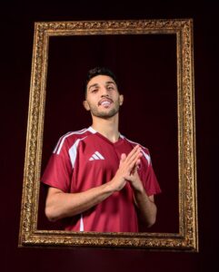 Man in a maroon Adidas jersey stands inside an ornate gold frame, hands pressed together in prayer against a dark burgundy background.