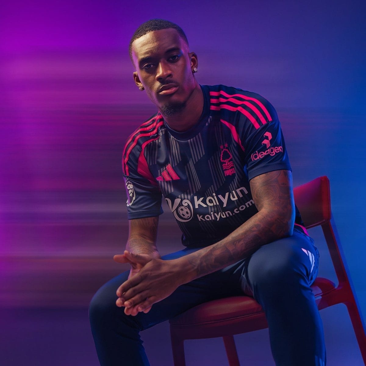 Portrait of a man in a dark pink and navy soccer jersey sitting on a red chair, hands folded, against a pink–purple gradient backdrop.