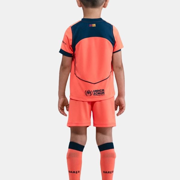 Back view of a child in a bright orange soccer kit with navy accents, UNHCR logo on the shorts, and neon cleats.