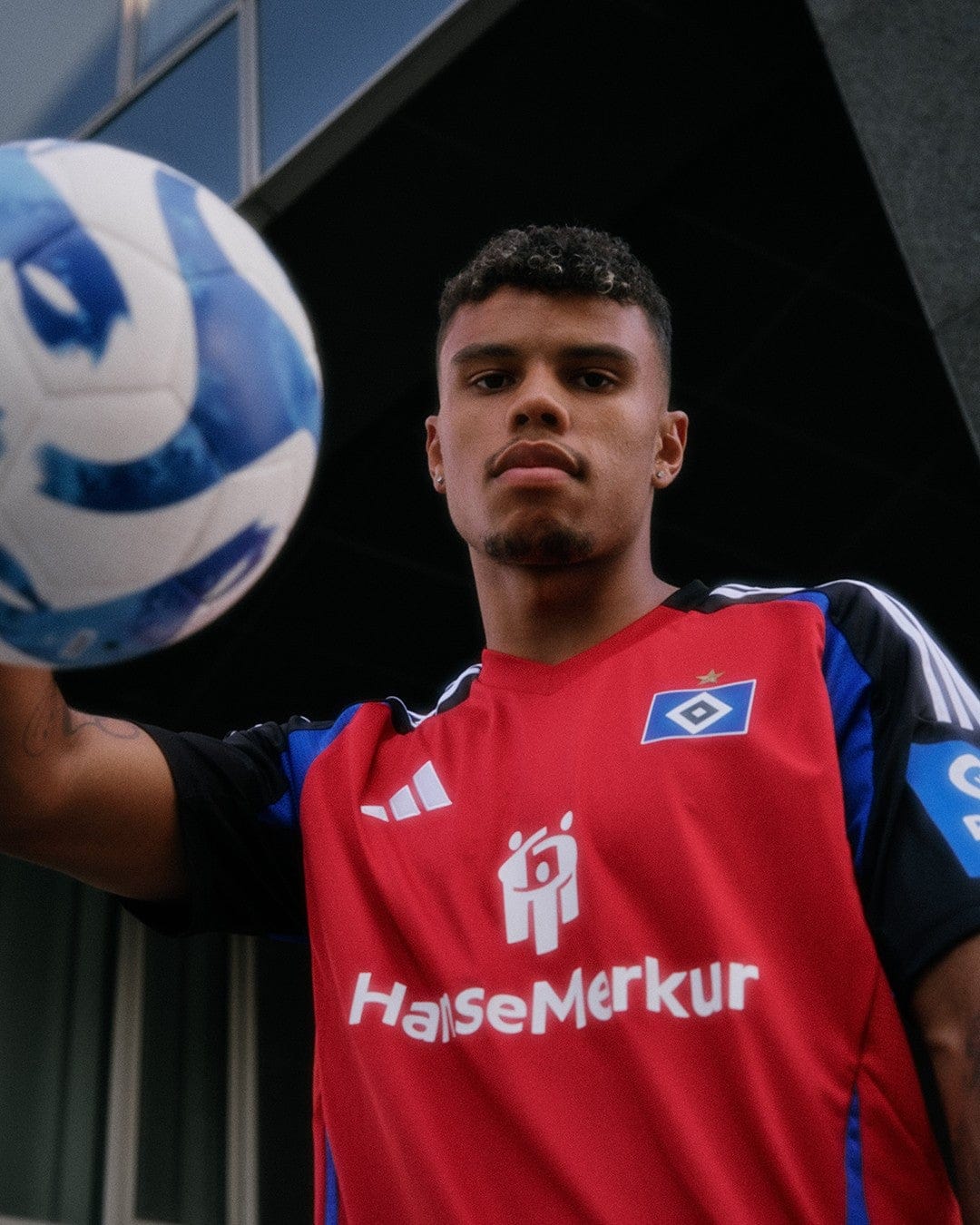 vvwlMr2mtu4MBmD | UAE SPORT STORE Young male soccer player in a red jersey with Hanse Merkur logo, ball hovering near his brow as he controls it outside a building.
