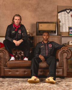 Two men sit on a brown leather sofa in a retro living room wearing black striped jerseys with crests and red scarves; vintage radio and framed jersey behind them.