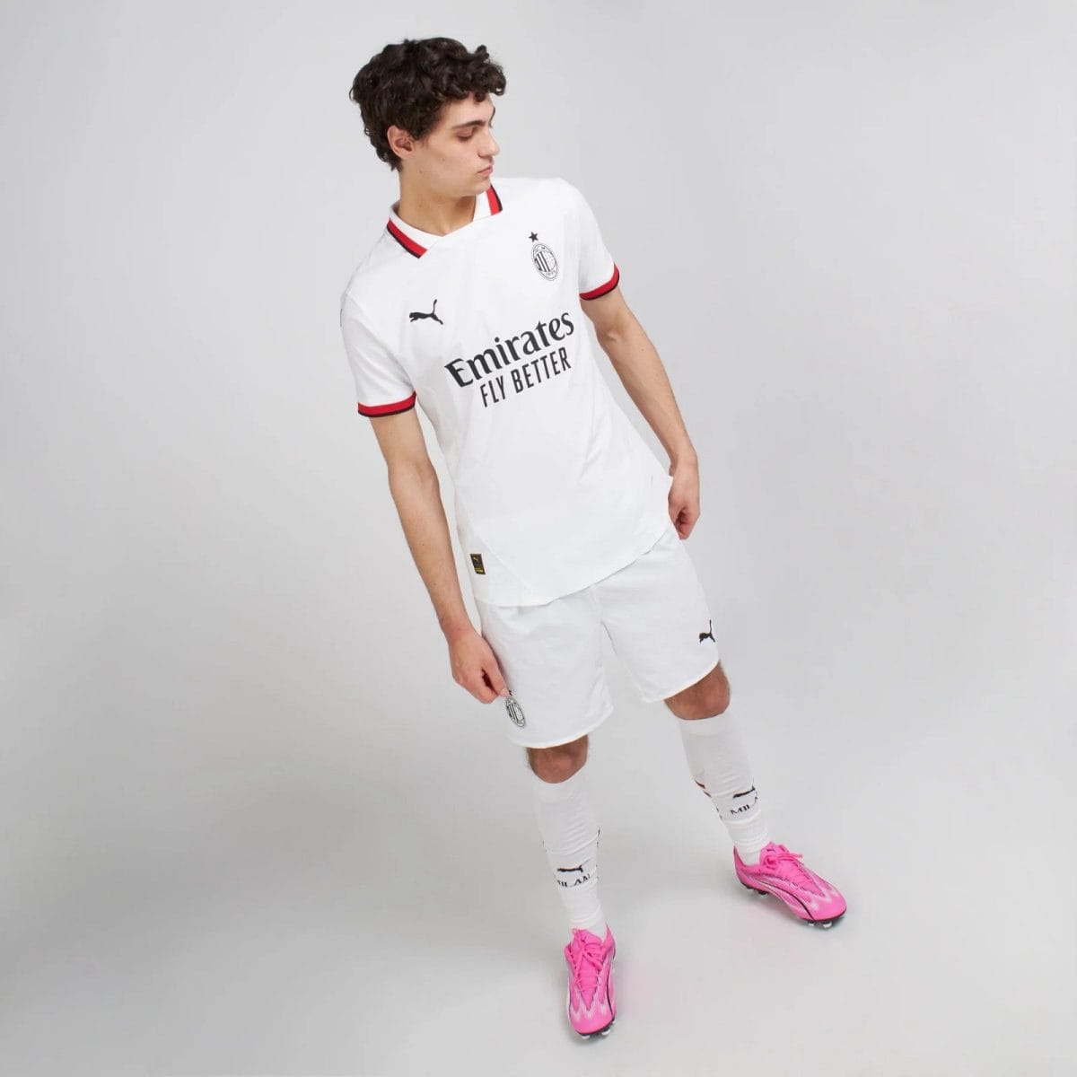 Soccer player posing in a white kit with red trim and pink cleats against a light gray background.
