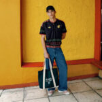 Young man in a black patterned shirt and jeans stands against a bright yellow wall, holding an Adidas tote bag.