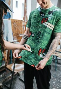 Two people in a studio create art on a green camouflage t-shirt with marker drawings, one holding the shirt while the other marks it.