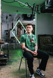 Portrait of a man in a green graphic T‑shirt sitting on a chair in an art studio, neon diamond frame behind him with painting supplies around.