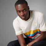 Portrait of a young man in a white sweater with rainbow stripes, seated against a gray backdrop and looking slightly to the side.