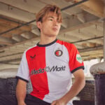 Man in a red-and-white striped soccer jersey sits against a concrete barrier, looking to the side.