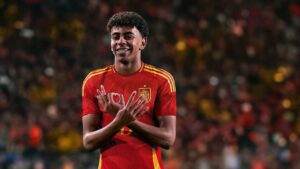 Young footballer in a red Spain national team jersey smiles while crossing his arms in front of his chest on a crowded stadium background.
