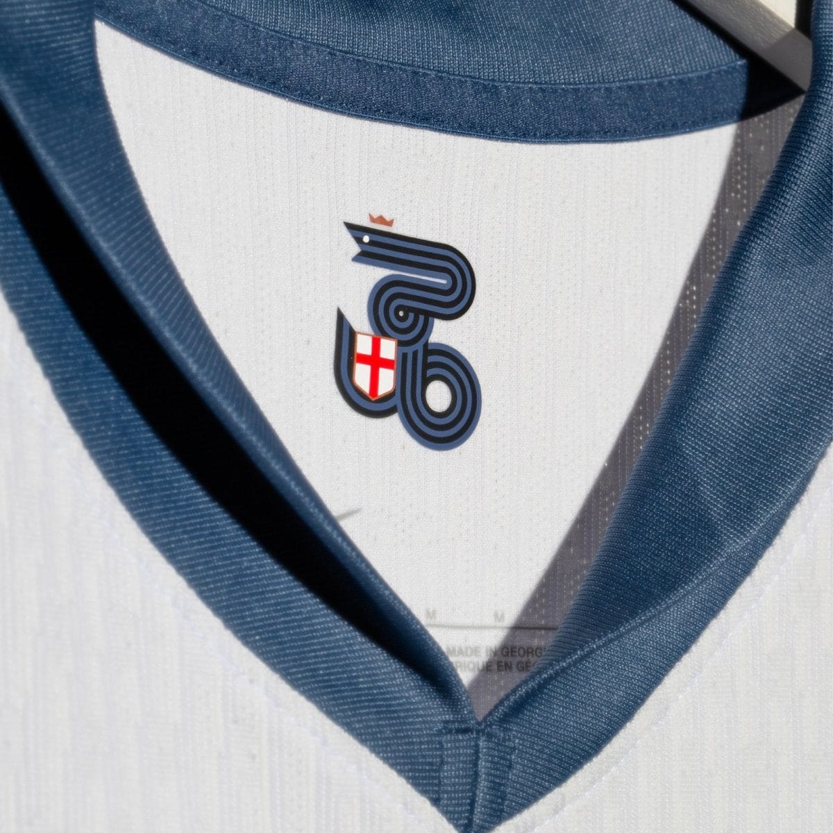 Close-up of a white shirt with a blue necktie, featuring a small crest: a navy stylized design with a tiny crown and a red cross shield.