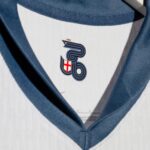Close-up of a white shirt with a blue necktie, featuring a small crest: a navy stylized design with a tiny crown and a red cross shield.