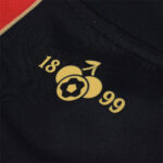 xNUuWCbqjEbBQAR | UAE SPORT STORE Close-up of a black fabric featuring a gold logo with the numbers 18 and 99 and a simple circular fruit motif.