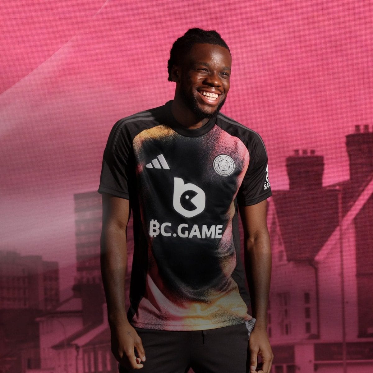 Smiling Black man wearing a BC.GAME football jersey against a pink cityscape backdrop.