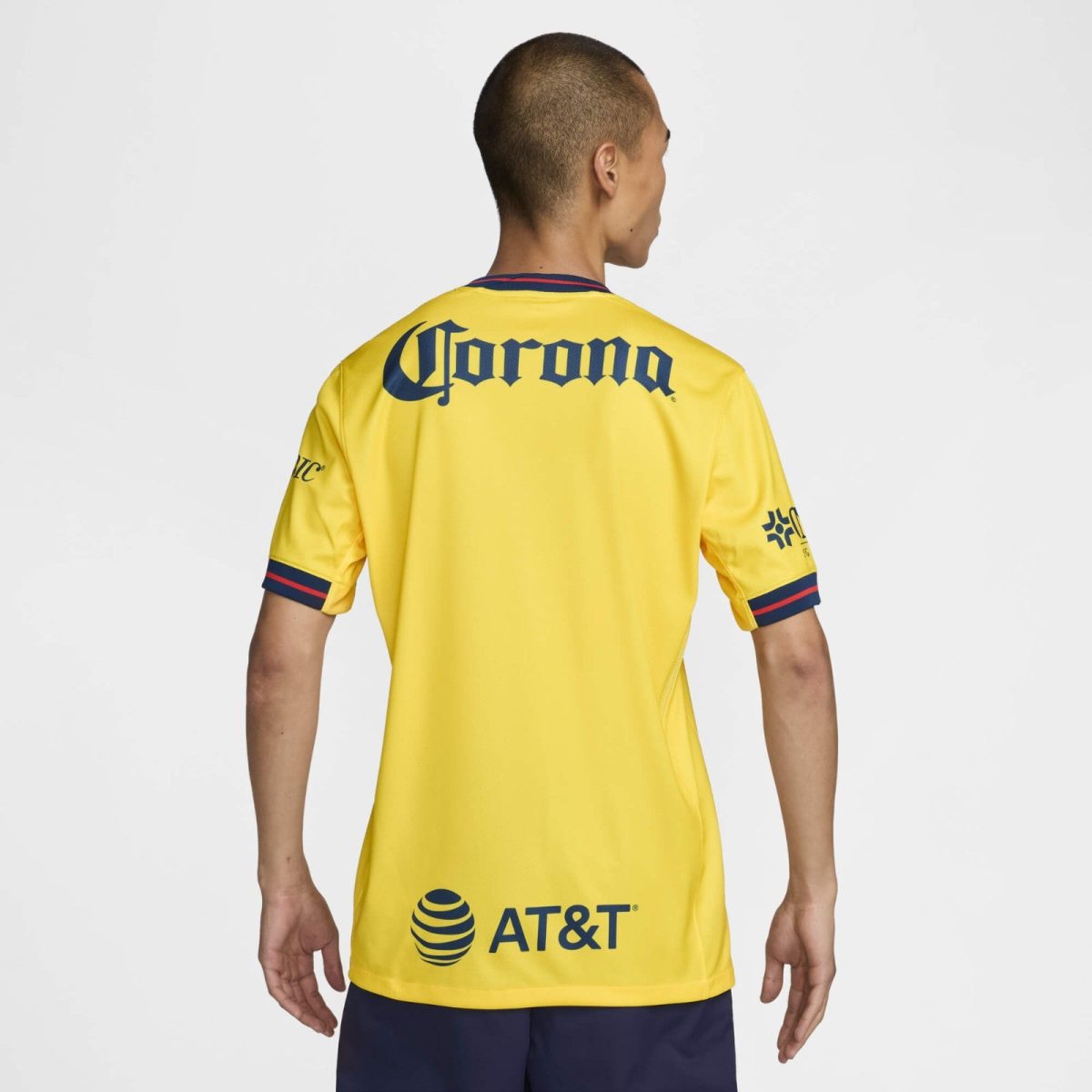 Back view of a bright yellow sports jersey with navy logos: Corona on upper back and AT&T toward the lower back, blue/red trim on sleeves and collar.