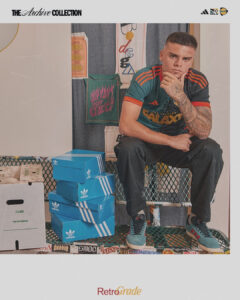 xfQxQlz9Ox3BPBs | UAE SPORT STORE A man sits on a metal crate wearing a teal and orange Adidas Galaxy jersey, surrounded by stacked blue Adidas shoe boxes and vintage banners in a studio setting.