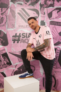 xv32B3Gw12IuUXi_4211bb27-cd98-4880-804c-65123b39f158 | UAE SPORT STORE Man in a pink striped jersey with tattoos on his arms, one foot resting on a white pedestal, posing against a pink collage backdrop