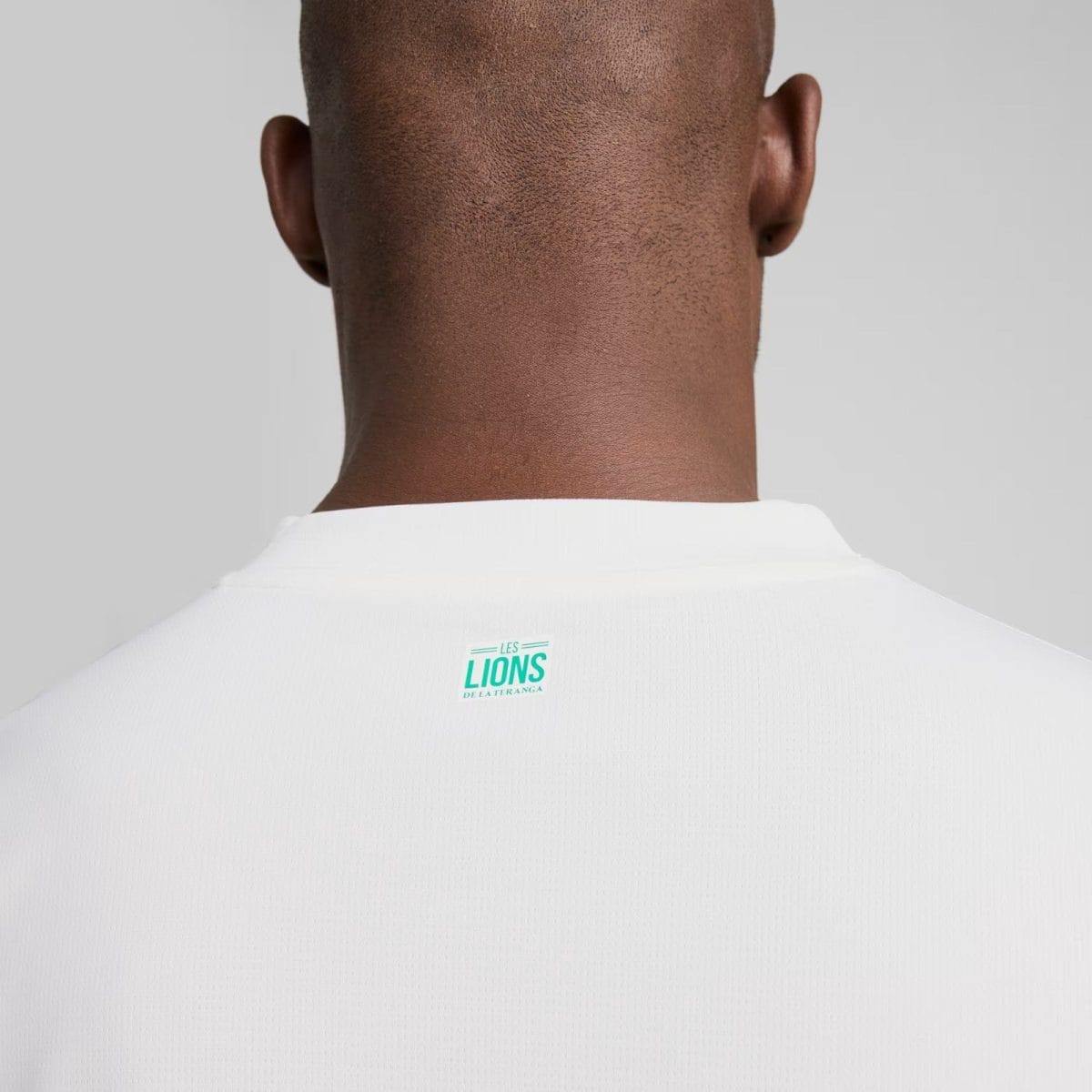 Back of a person wearing a white sports jersey; a small turquoise 'LES LIONS DE LA TERANGA' logo is on the upper back.