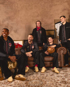 Five teammates in matching black striped jackets sit and stand on a vintage brown sofa in a retro living room, smiling at the camera.
