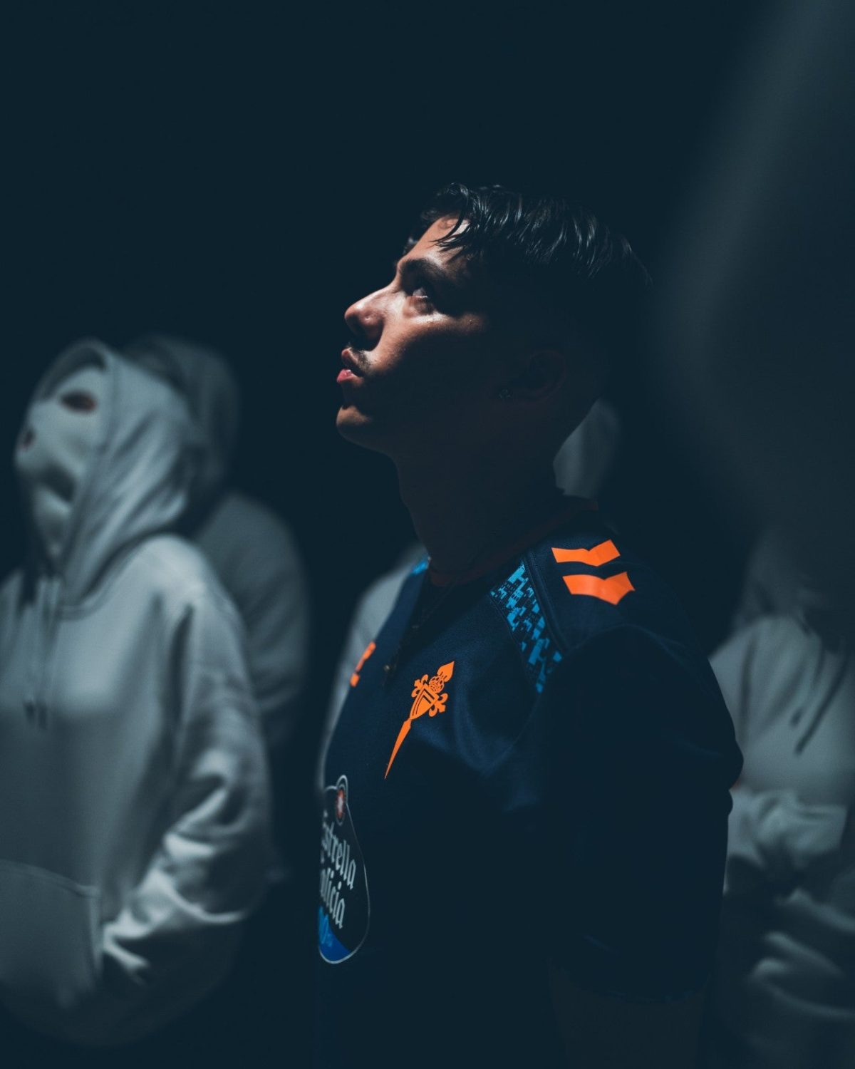 Side profile of a man in a dark blue jersey with orange details, looking upward in a dim room with blurred figures in the background.