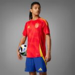 Soccer player in a red jersey with Spain crest holding a ball against a gray studio background.