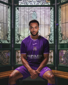 Portrait of a male footballer seated on a wooden bench, wearing a purple jersey with Napoleon Sports & Casino logo, in a stained glass backdrop.