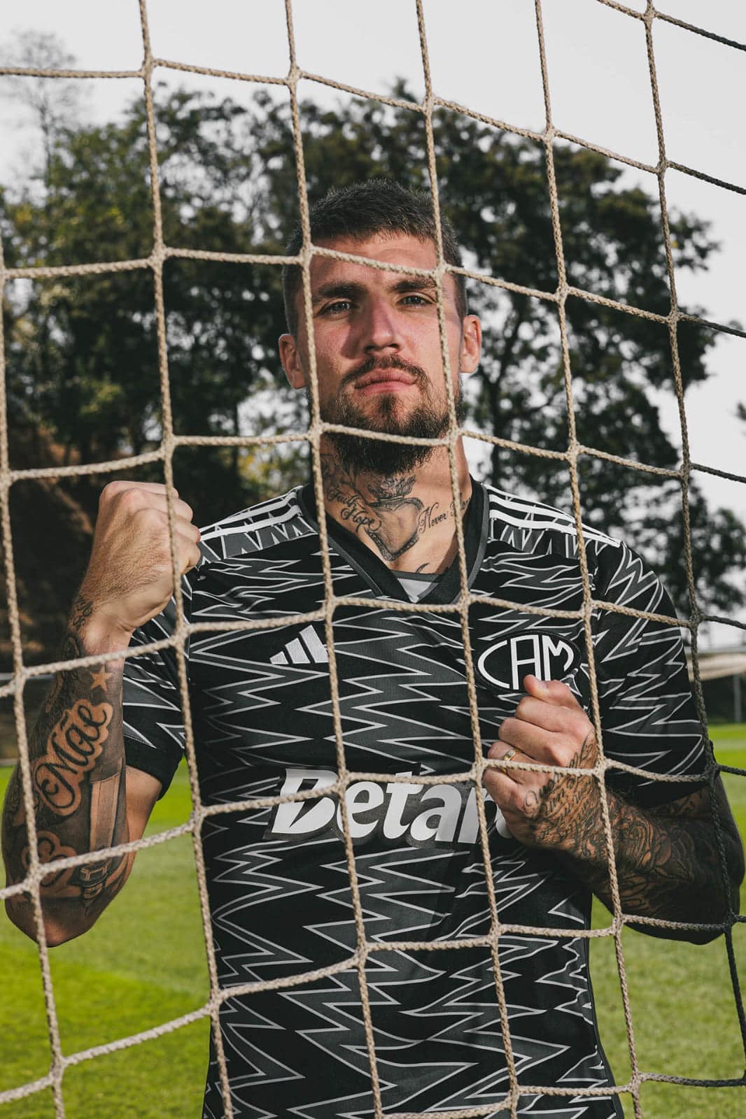 yblak1pBvRX9SdC | UAE SPORT STORE Tattooed man standing behind a soccer goal net, wearing a black zigzag-pattern jersey and looking at the camera.