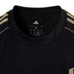 yi | UAE SPORT STORE Close-up of a black shirt collar with gold stripes along the shoulders and an Adidas logo visible on the interior label (size M).