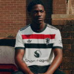 Young man sitting outdoors against a brick wall, wearing a white striped t‑shirt with red, black, and gray bands and logos.