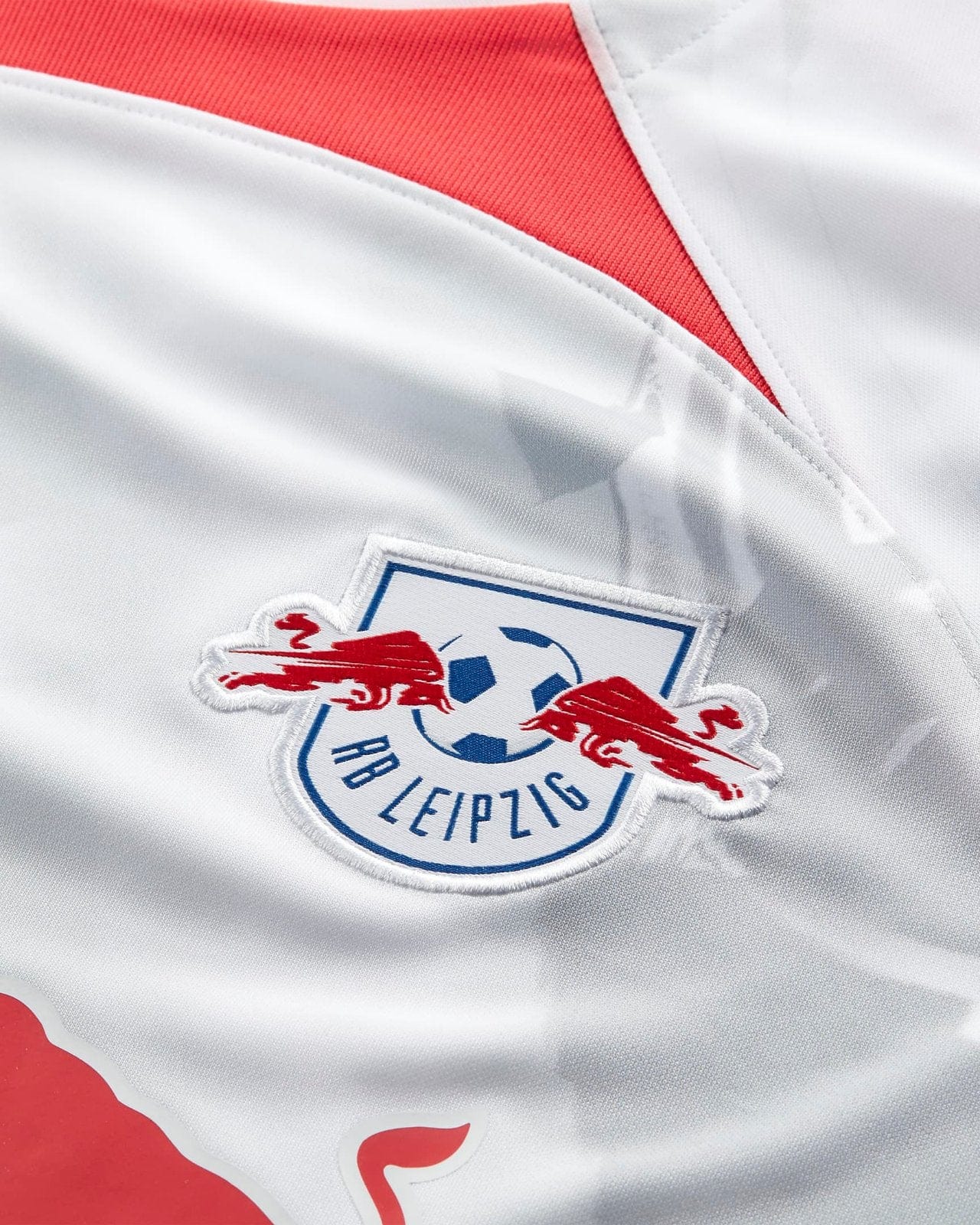yr4cBLaZxozRZvw | UAE SPORT STORE Close-up of a white RB Leipzig jersey with a blue-edged club badge and red trim on the collar area.