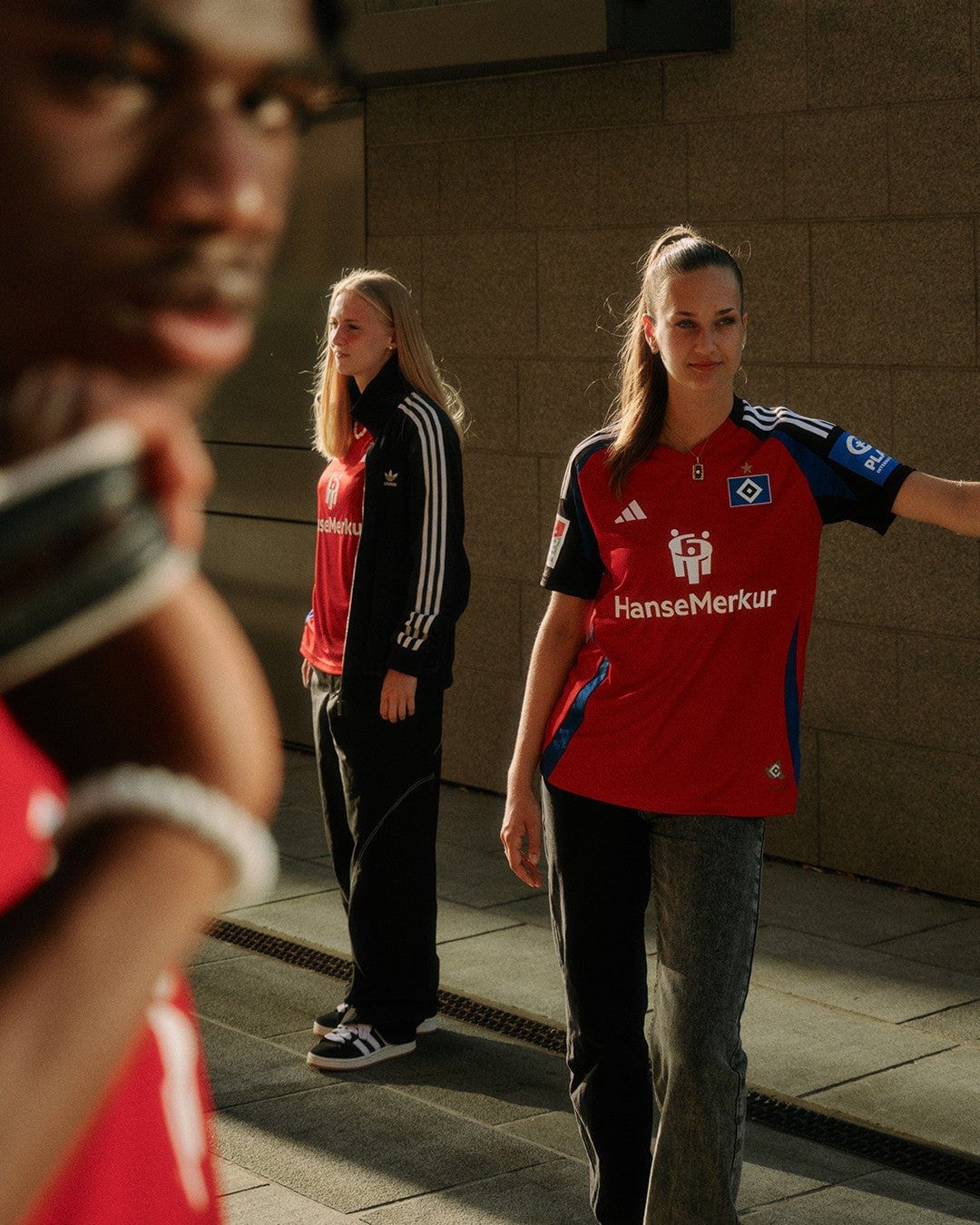 yxrDcNY9uojkGwt | UAE SPORT STORE Three young women in red football jerseys stand near a stone wall, sunlight glowing on one as she smiles slightly; another in a black Adidas jacket stands behind.