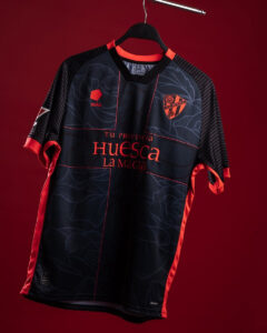 Dark blue and red Huesca football jersey hanging on a hanger against a red background, showing sponsor text 'Huesca La Magia' and crest.