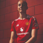 Person wearing a red Manchester United football jersey, posing against a brick wall; sponsor logos visible on chest and sleeve.
