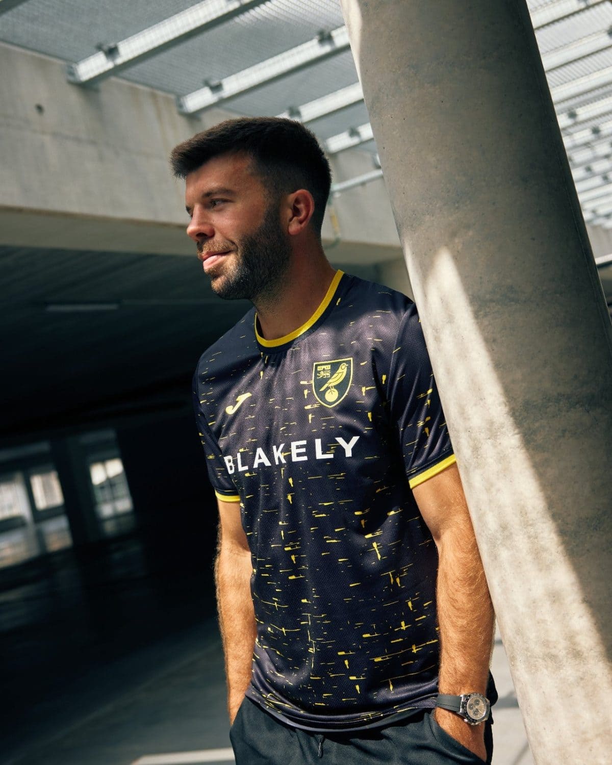 Man in a dark blue Blakely jersey with yellow specks leans against a concrete pillar in an indoor setting.