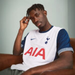 Young man wearing a white and blue Tottenham jersey sits on a brown couch, resting his head on his hand.