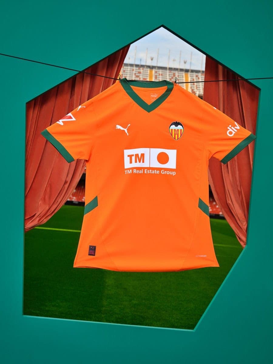zMipBcRIzLuRgaC | UAE SPORT STORE Valencia CF orange soccer jersey with green trim hanging on a clothesline inside a teal geometric frame, sponsor TM Real Estate Group visible.
