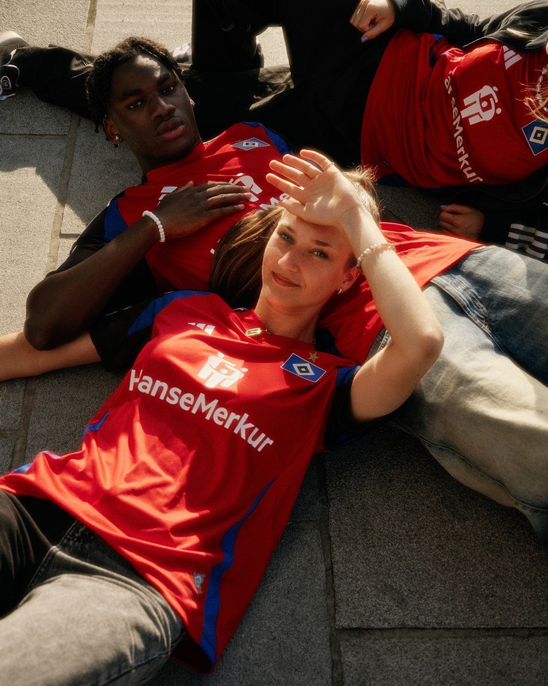 zSm2ll08ZAmXfRR | UAE SPORT STORE Two young people in red team jerseys rest on stone steps, the woman in front smiles and shields her eyes with a hand.