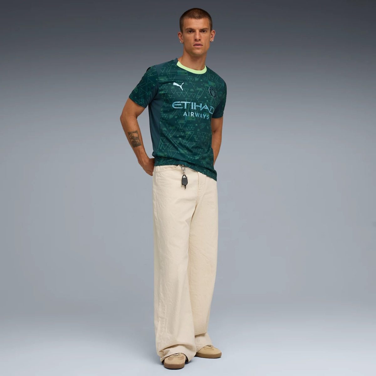 Male model wearing a teal Manchester City jersey with Etihad Airways logo and beige wide-leg pants, standing with hands on hips against a gray backdrop.