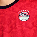 Close-up of a red football jersey featuring the Egyptian FA crest with a soccer ball and green Arabic text, above a row of white stars near the chest area.