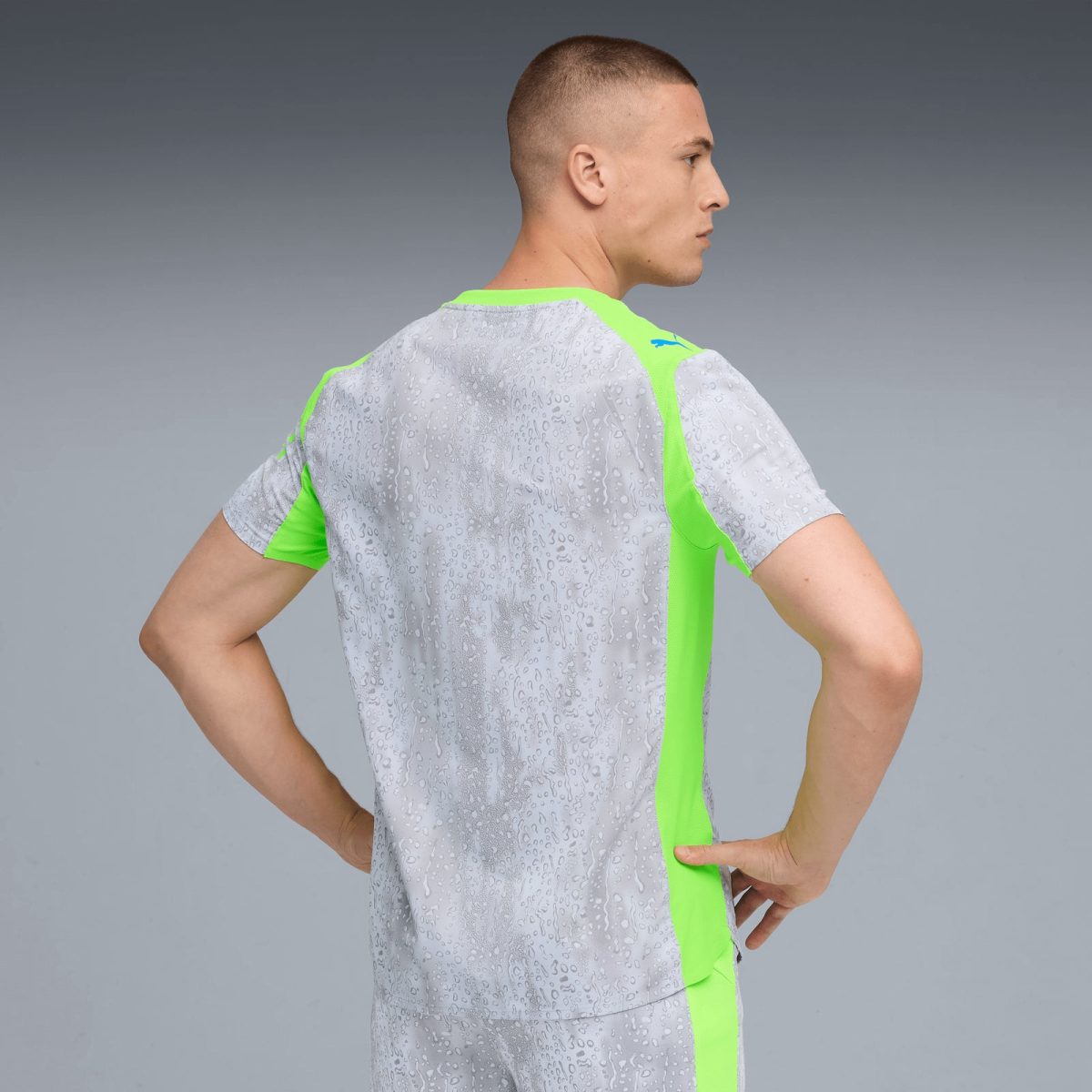 Side profile of a man wearing a light gray athletic shirt with neon green panels, standing with hands on hips against a gray gradient background.