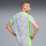 Side profile of a man wearing a light gray athletic shirt with neon green panels, standing with hands on hips against a gray gradient background.