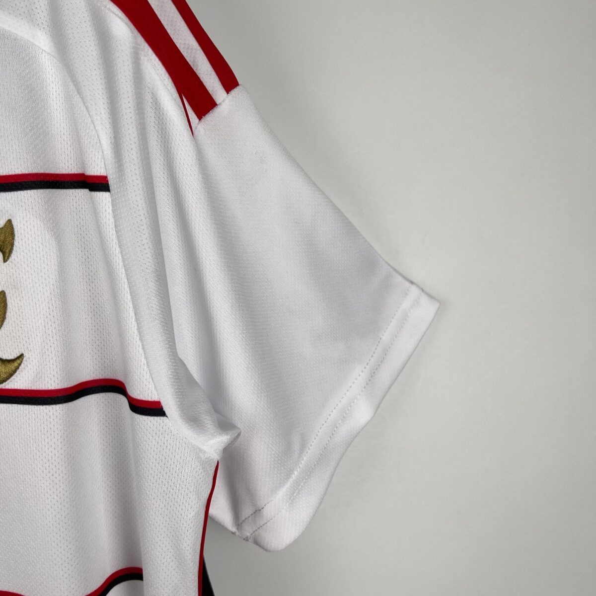 White football jersey with red and black stripes and gold emblem on chest.