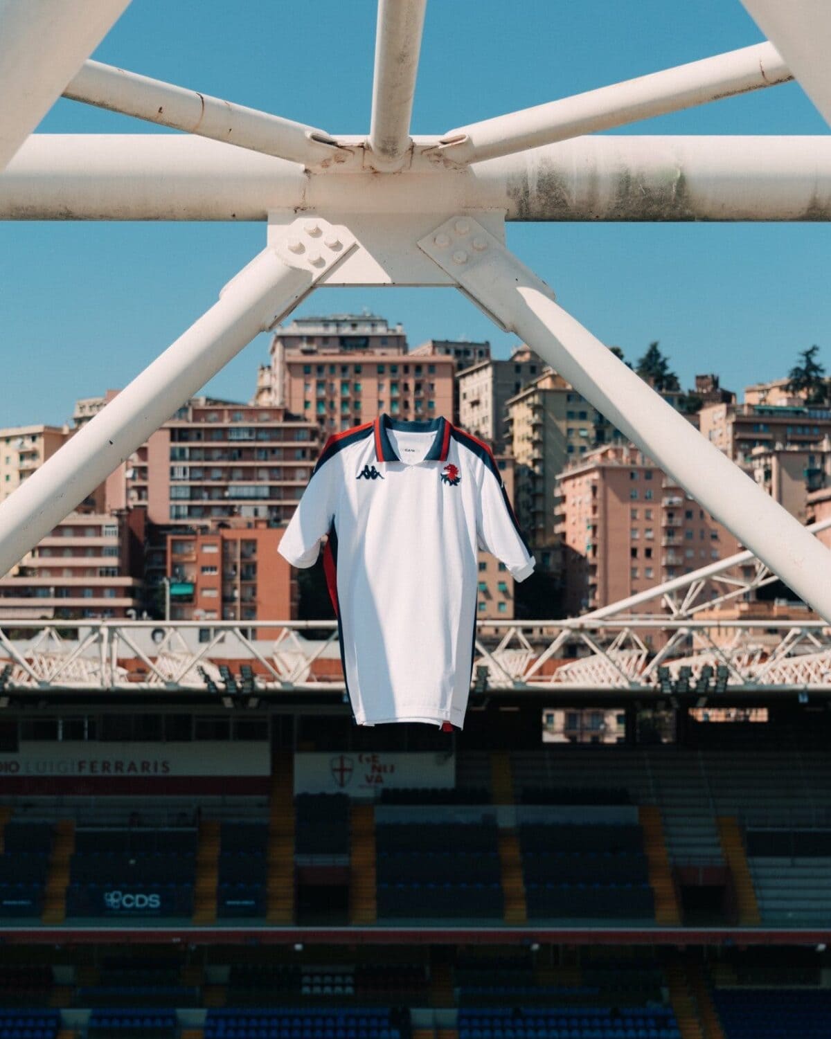 Genoa home football jersey 2024/25 hanging on stadium structure with city background.