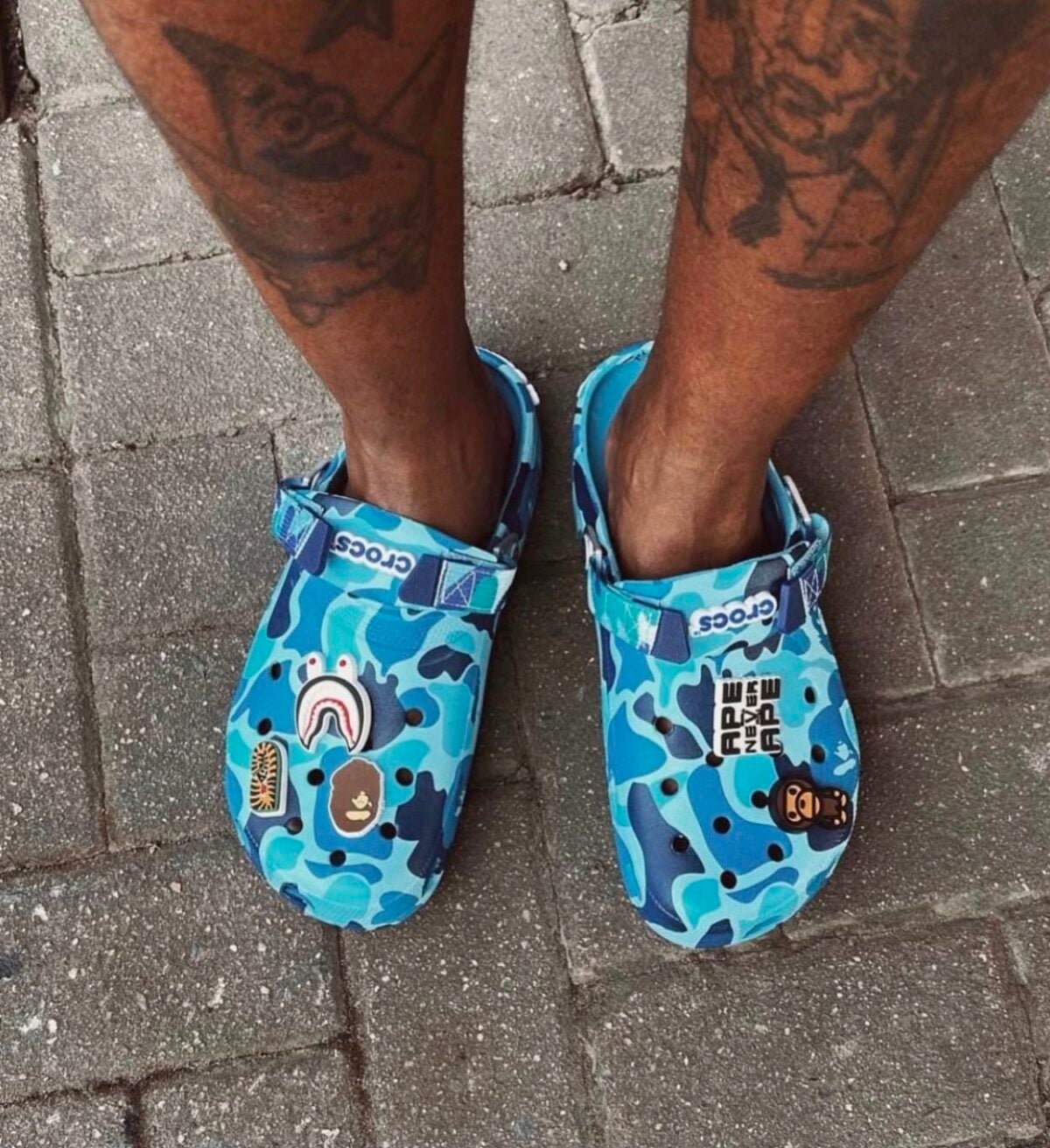 Blue camouflage Crocs with cartoon patches on a person's feet on pavement.