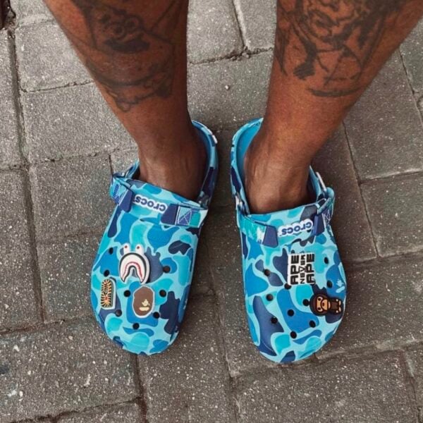 22310e70-01c5-48c7-9f92-0bfda06093e8 - UAE SPORT STORE Blue camouflage Crocs with cartoon patches on a person's feet on pavement.
