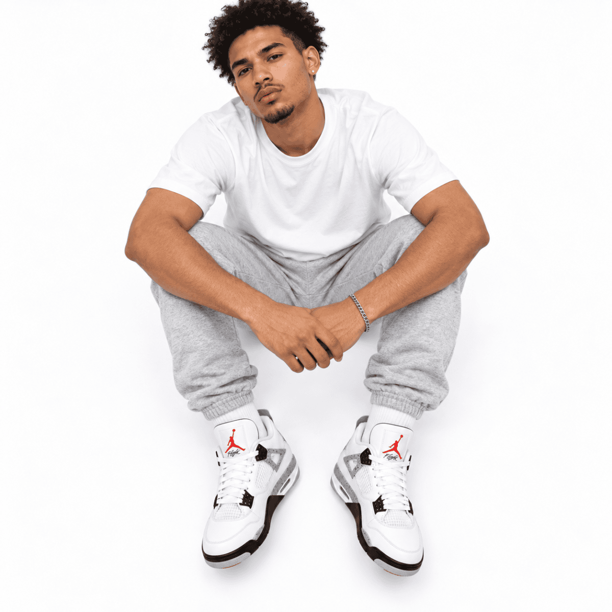 Young man in white t-shirt, gray sweatpants, and white sneakers sitting on floor.