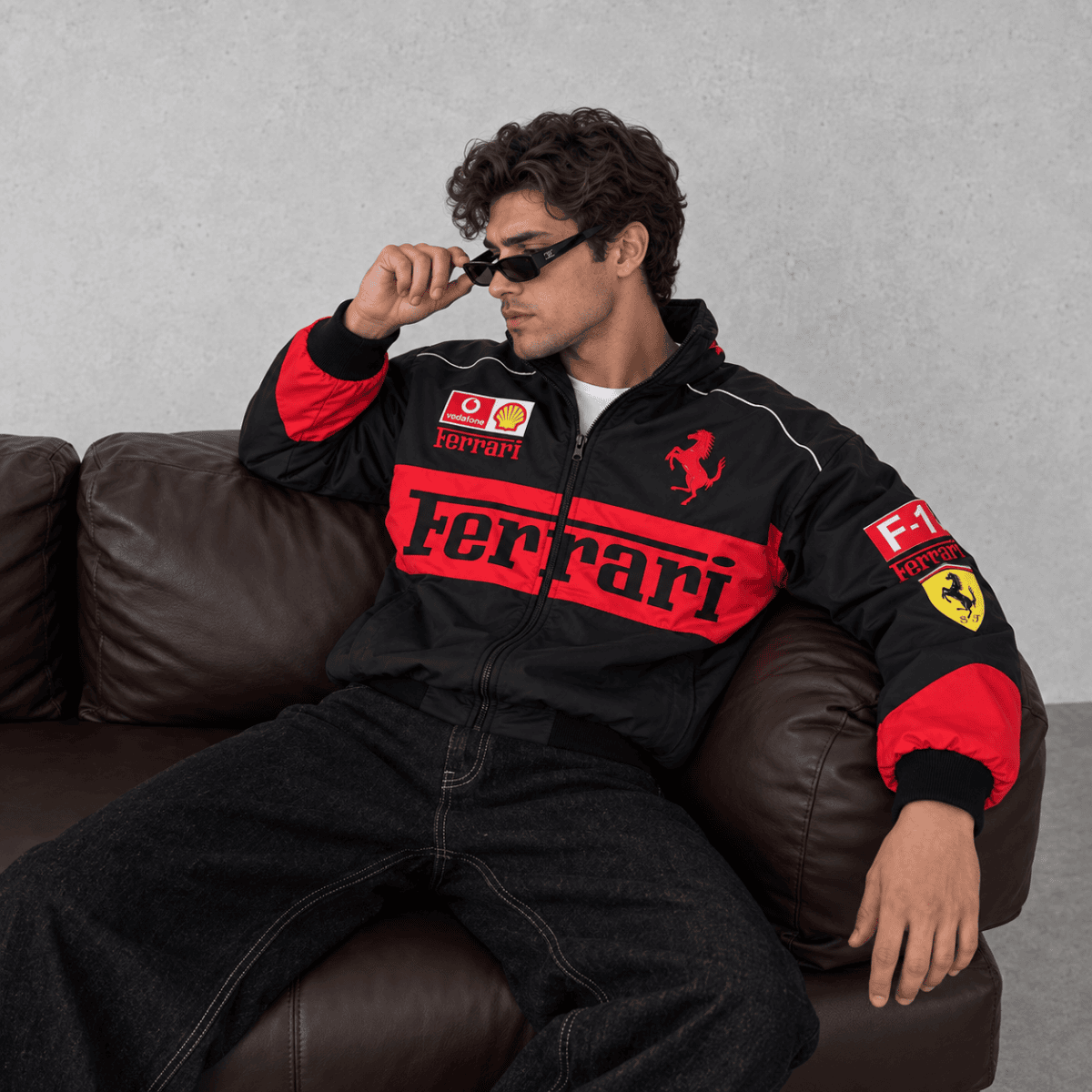 Man wearing Ferrari Formula 1 team jacket with logos, sitting on a brown sofa.