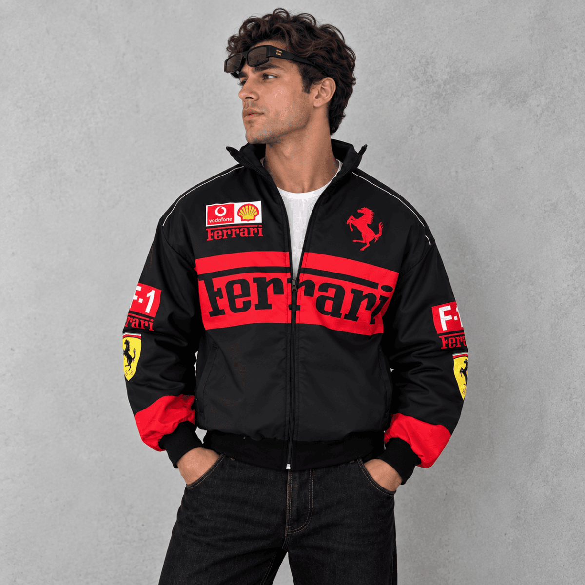 Man wearing black Ferrari racing jacket with red logo and patches, standing against gray background.